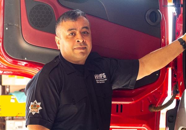 A male firefighter stands in front of the door to a fire engine, leaning on the seat, wearing the new uniform shirt.