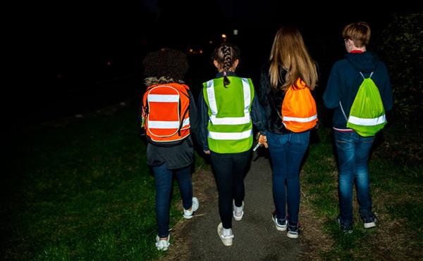 The back of four school-age children walking in the dark with high visibility strips on jackets and bags they are wearing.