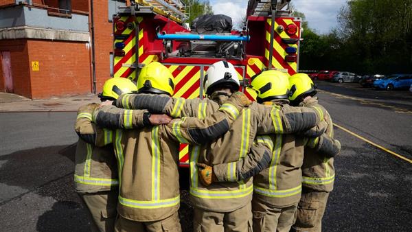 Five firefighters with their arms around each other stand together in front of a fire engine.
