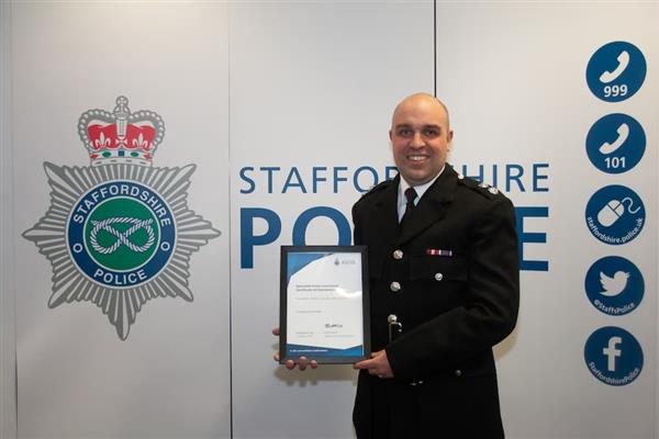 Watch Commander Huckle in full uniform stood in front of a logo of Staffordshire Police holding a certificate.