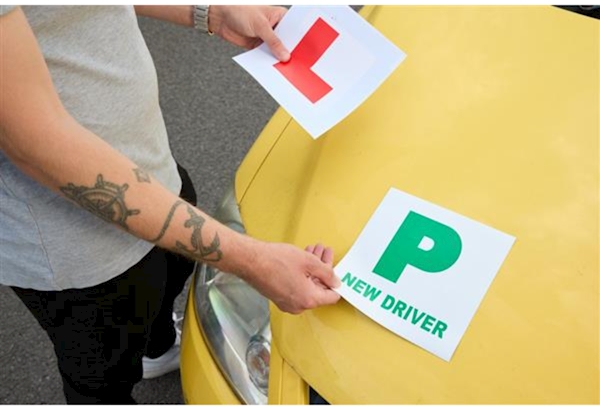 A green 'P' plate and a red 'L' plate being held on a yellow car's bonnet by a young man
