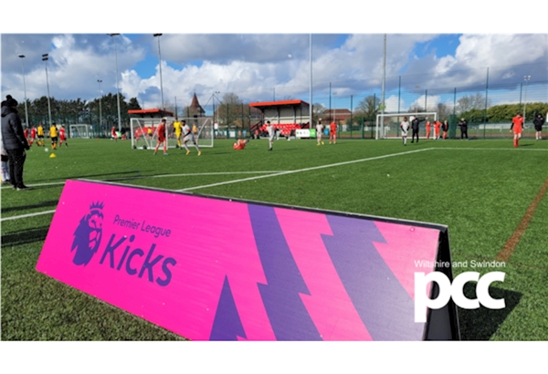 Premier league kicks sign in foreground with training session at Foundation Park, Swindon in background