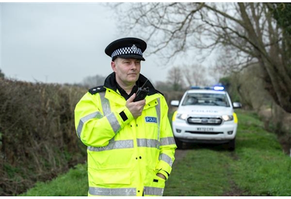 A British police inspector in a high-visibility jacket holding a radio, with a police car in the background.