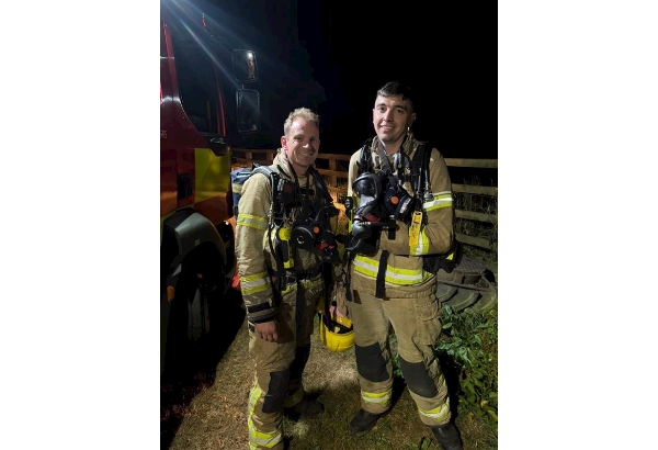 Two on-call firefighters stood smiling, wearing breathing apparatus next to a fire engine