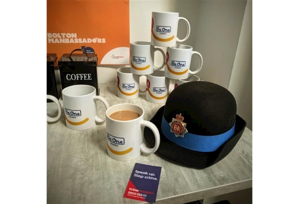 Image Showing PCSO Hat, Be One Homes Cups and Bolton Manbassadors Merchandise, Crimestoppers Cards and a mug of Tea