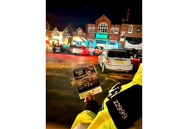 A police officer holds a "home and dry" leaflet in front of a brightly lit town centre at night